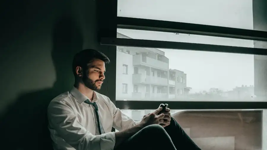 A man sitting by a window, reading messages on his phone